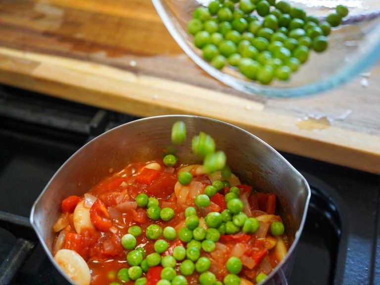 Peas being poured into a pan with cooked food, tomato beans and peppers